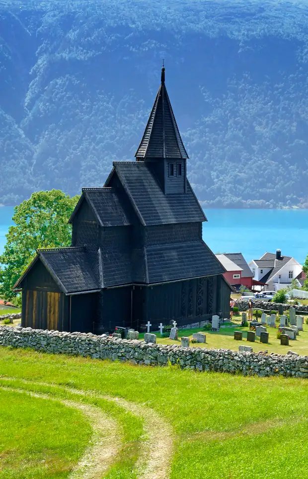 Urnes stavkyrkje i mørkt, tjærebredd tre, med steingjerde og Lustrafjorden bak.